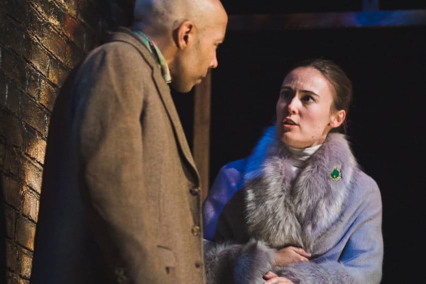 A production image from ‘Sherlock Holmes – The Hunt for Moriarty.’ A man wearing a long brown coat listens intently as a woman wearing a lilac furry coat speaks to him.