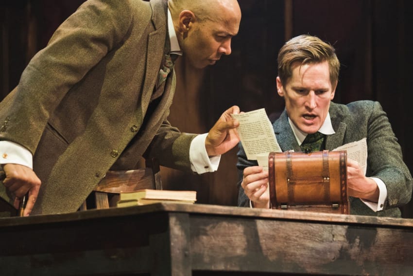 A production image from ‘Sherlock Holmes – The Hunt for Moriarty.’ A man sits at a desk, with another man stood up, leaning over his shoulder. They look intrigued and shocked by a piece of paper which they have taken out of a small wooden box.