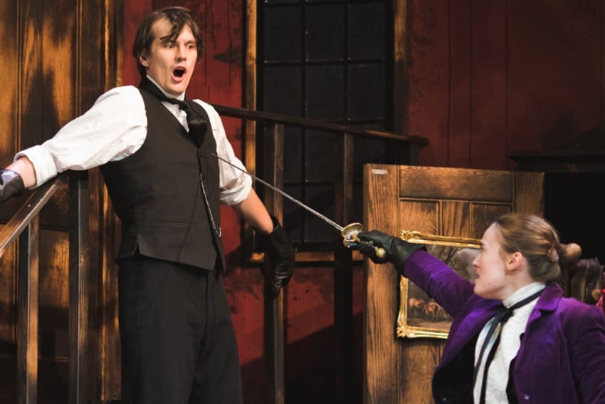 A production image from ‘Sherlock Holmes – The Hunt for Moriarty.’ A woman wearing a purple blazer draws a sword at a man, who looks shocked as he leans back against a staircase.