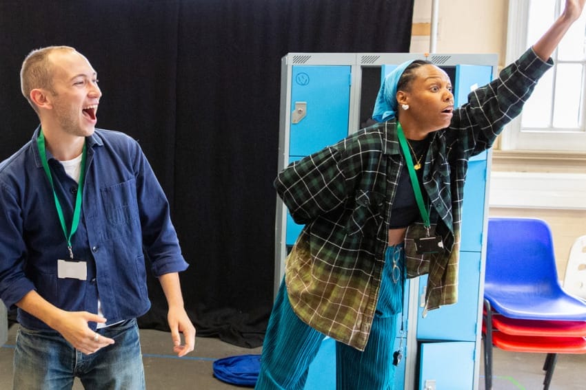 Isaac and Chileya in rehearsals – Isaac smiles whilst Chileya waves at something in the distance.
