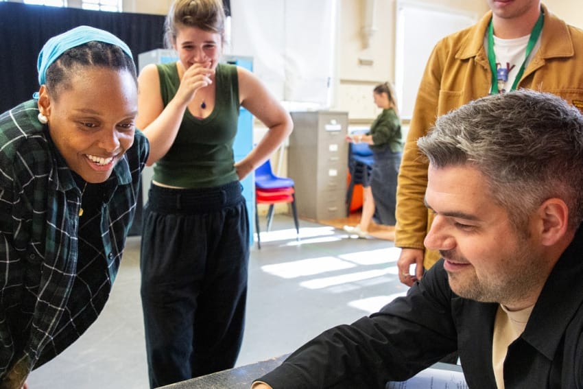 Actors and director in rehearsals – Jake shows something on his phone to the three actors who stand around the table watching.
