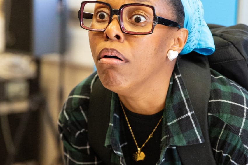 Chileya in rehearsals – she’s wearing big brown glasses and a green flannel shirt and is holding an open notebook. She is making a funny face and holding down on the paper of the book.