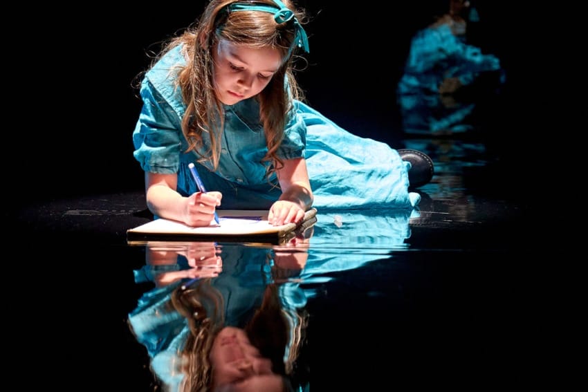 A production image from ‘Don’t Look Now.’ A young girl wearing a light blue dress is laying on the floor, drawing a picture on a piece of paper. He reflection can be seen on the shiny black floor.
