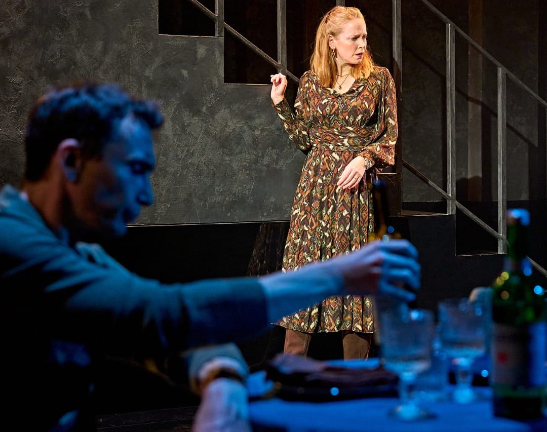 A production image from ‘Don’t Look Now.’ John, blurred in the foreground, is sat at a restaurant table, picking up a bottle. In the background, Laura stands next to some stairs. She has a confused look on her face, as if she has just been told something shocking.