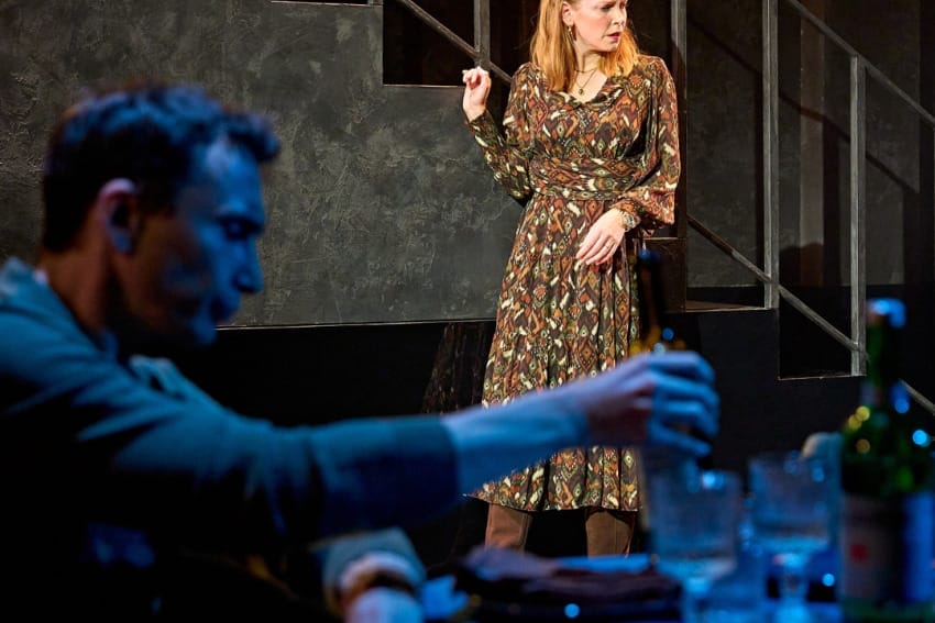 A production image from ‘Don’t Look Now.’ John, blurred in the foreground, is sat at a restaurant table, picking up a bottle. In the background, Laura stands next to some stairs. She has a confused look on her face, as if she has just been told something shocking.