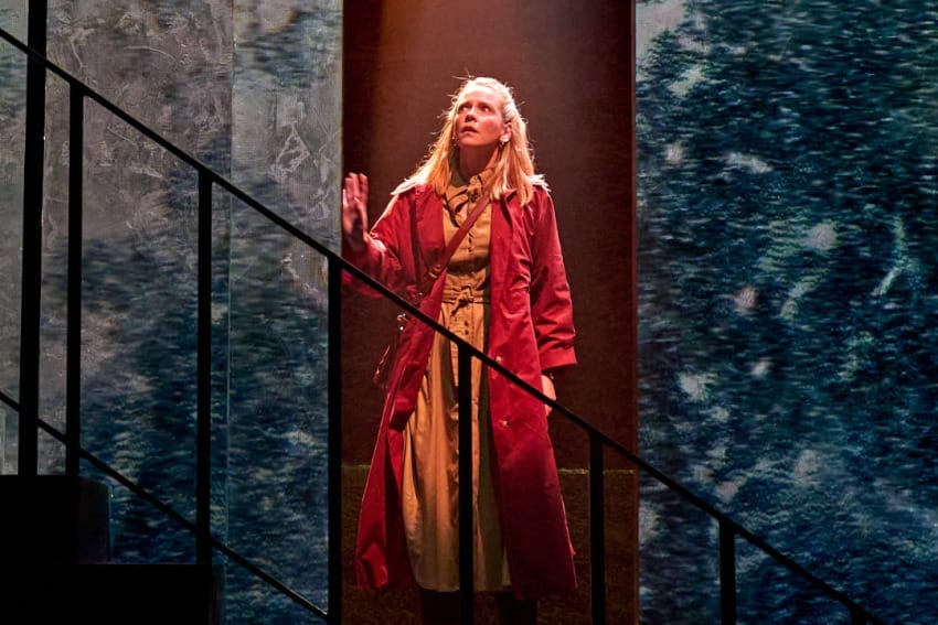 A production image from ‘Don’t Look Now.’ Laura stands in a doorway, with one hand resting on the door frame. She is looking up towards the sky, nervously. She is wearing a long, bright red coat.