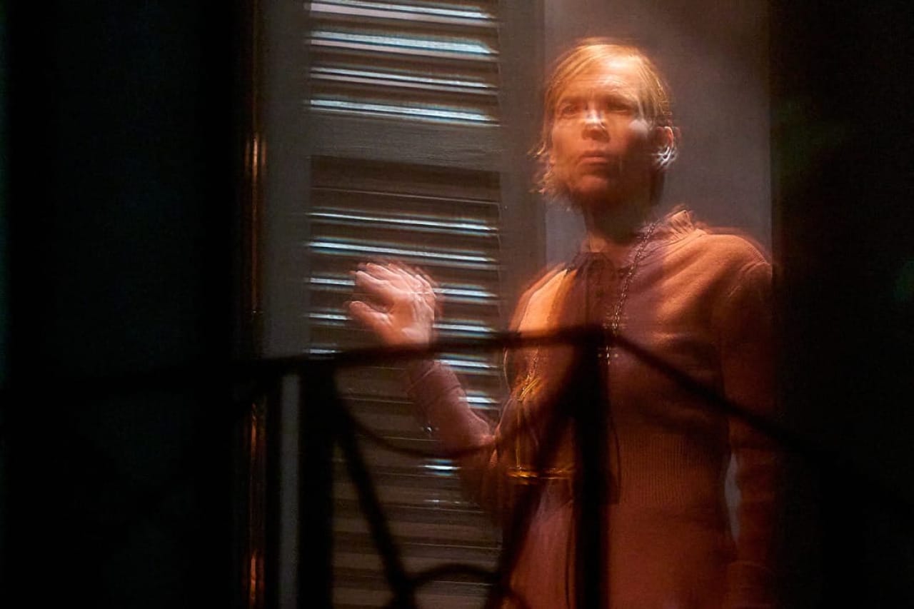 A production image from ‘Don’t Look Now.’ One of the sisters stands at the top of some stairs in the middle of a doorway, looking out towards the stage. The image has a blurred effect to it.