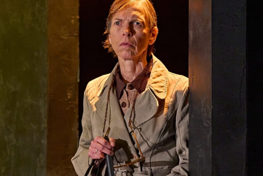 A production image from ‘Don’t Look Now.’ One of the sisters stands in a doorway, with a glowing yellow light shining from above her. She is holding a walking stick in her hands and is looking towards the centre of the stage, listening intently.