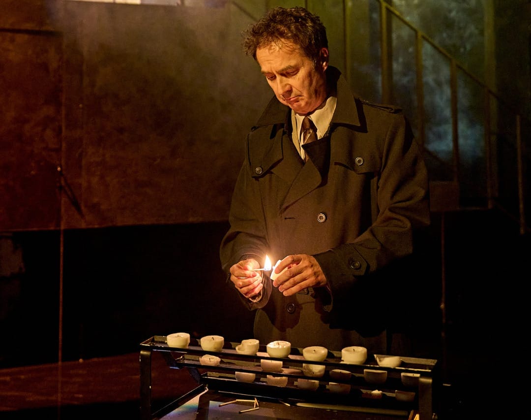 A production image from ‘Don’t Look Now.’ A man is standing behind a table with stacks of white candles on it. He has picked up one of them and is lighting it with a match.