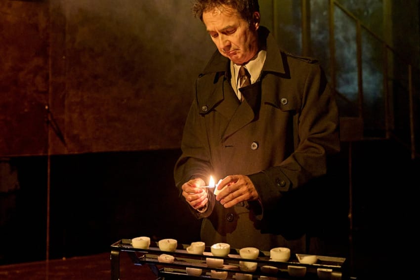 A production image from ‘Don’t Look Now.’ A man is standing behind a table with stacks of white candles on it. He has picked up one of them and is lighting it with a match.