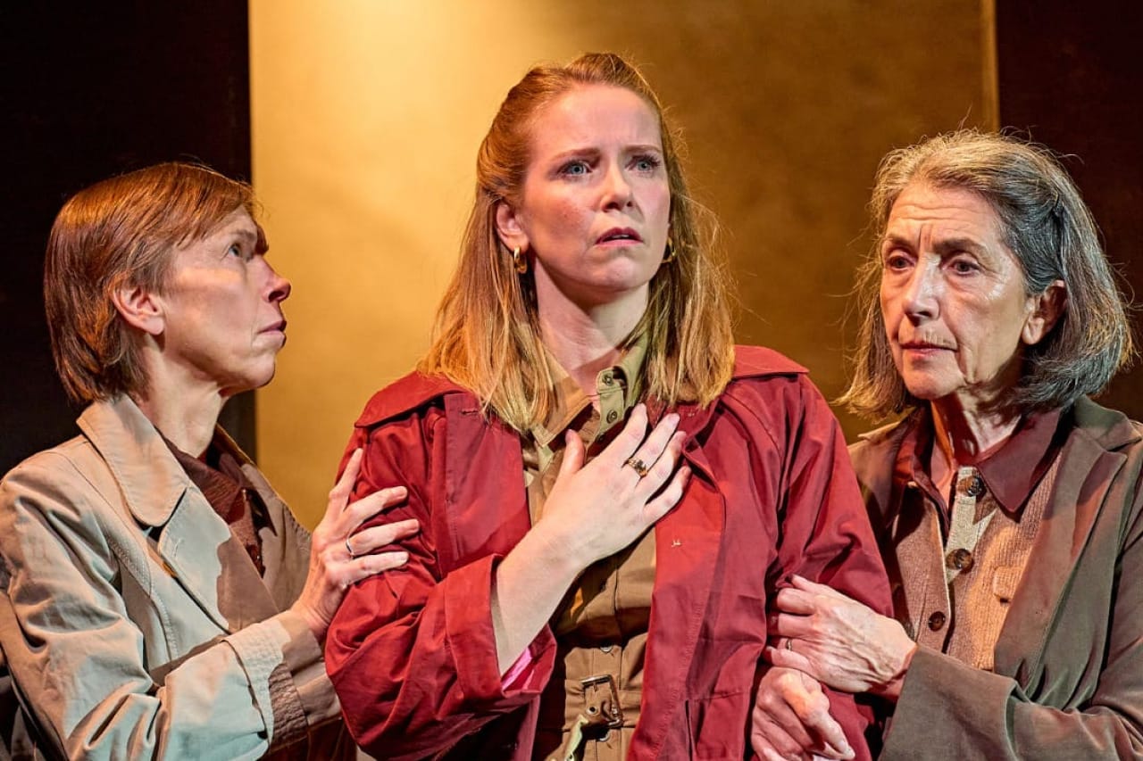 A production image from ‘Don’t Look Now.’ Laura stands in the middle of the two sisters, who are each clutching onto one of Laura’s arms. Laura holds one hand to her chest and looks out into the distance, with a look of concern on her face.