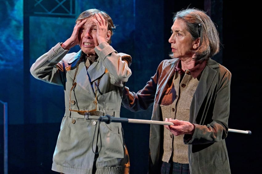 A production image from ‘Don’t Look Now.’ The two sisters stand together. Sister 1, on the right, places her hand on the shoulder of Sister 2, who is holding both hands to her head and has an extremely panicked expression on her face. The stage behind them is lit a deep blue colour.
