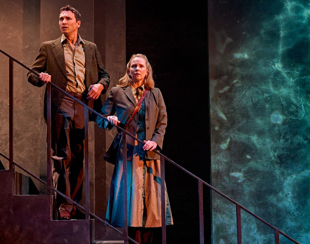 A production image from ‘Don’t Look Now.’ John and Laura stand half-way down some stairs, holding onto the railings. They both have slightly stunned looks on their faces, as if they have been spooked by a sight or a sound.