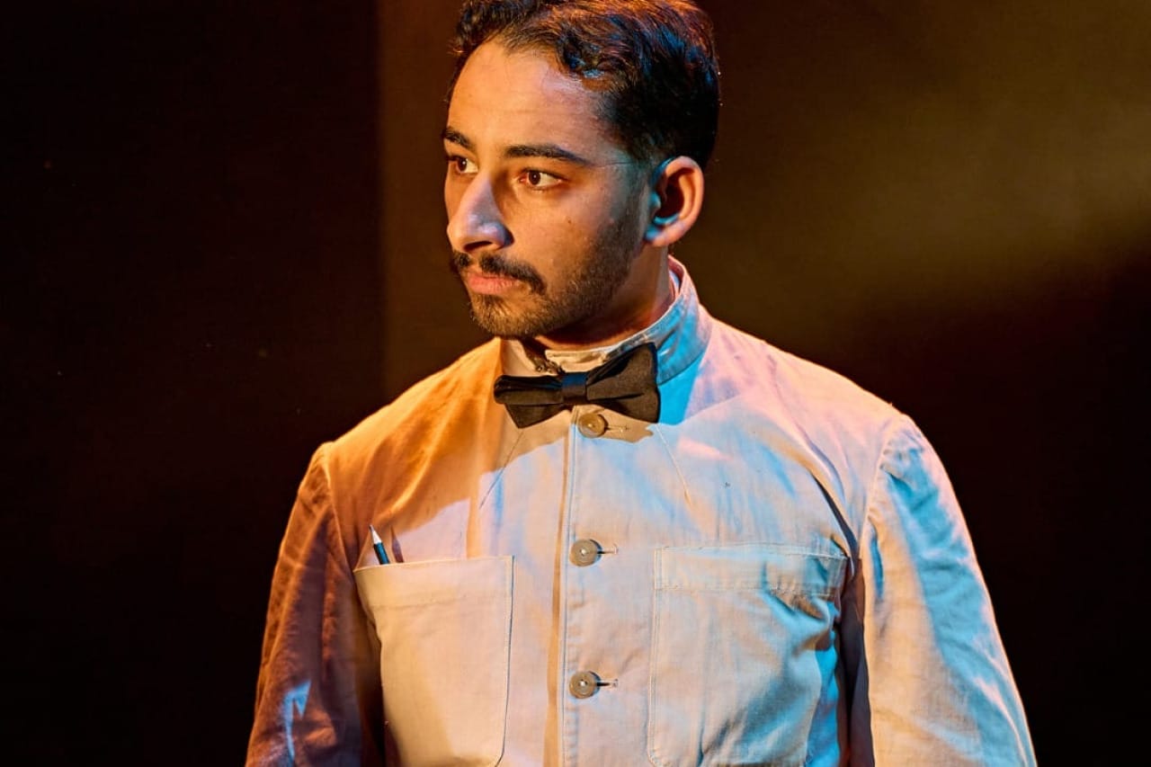 A production image from ‘Don’t Look Now.’ A restaurant waiter stands, wearing a smart white shirt and black bow tie. He is listening intently to something being said by another character.