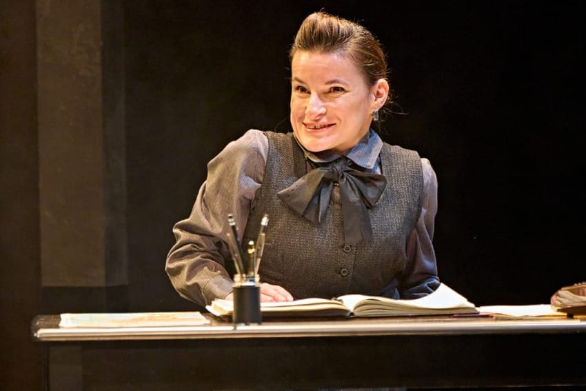 A production image from ‘Don’t Look Now.’ The hotel Clerk, wearing a smart black uniform, smiles as she stands behind a desk.