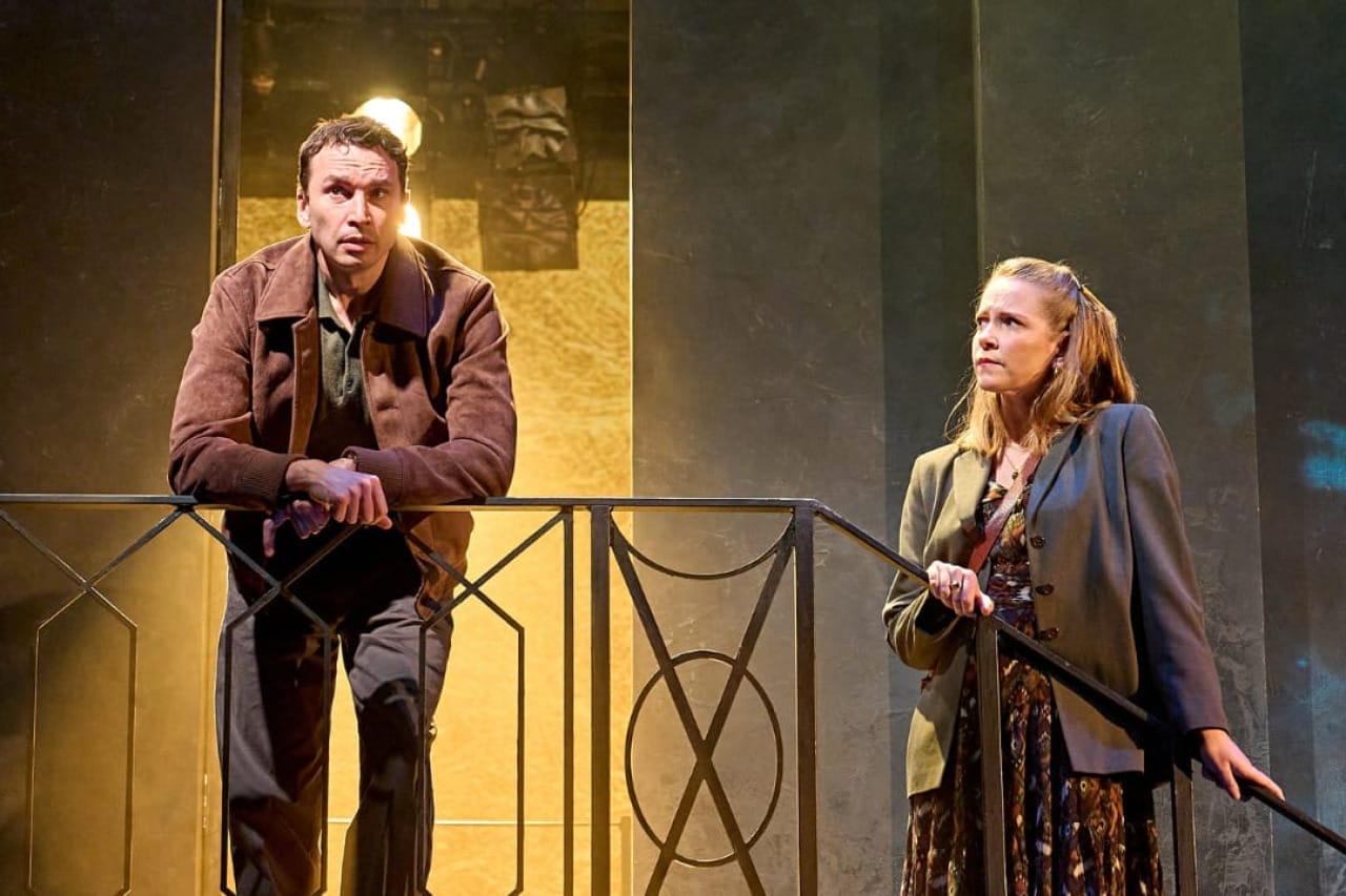 A production image from ‘Don’t Look Now.’ Laura stands half-way down some stairs, holding onto the railings, whilst John is at the top of them. Laura looks at John, worried. A glowing yellow light illuminates John from behind, from a doorway he is standing in front of.